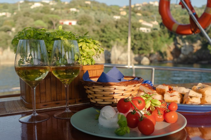 Two wine glasses, basil plant, tomatoes, bread on a table with scenic background.