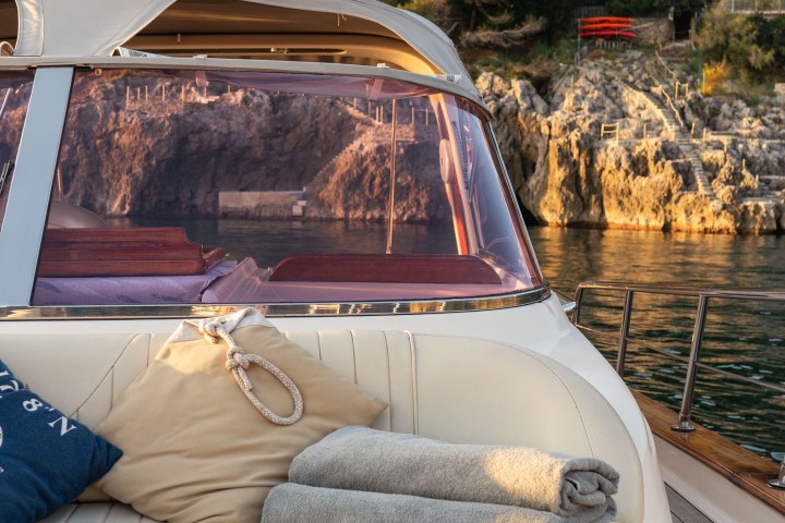 Boat deck with cushions and towels, overlooking rocky coastline and trees.