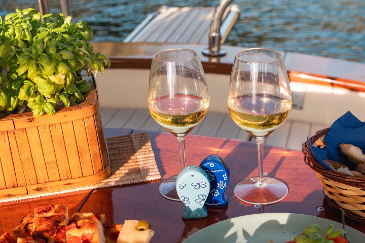 Two glasses of wine and plates of bread and tomatoes on a boat with water and rocky shore in the background.