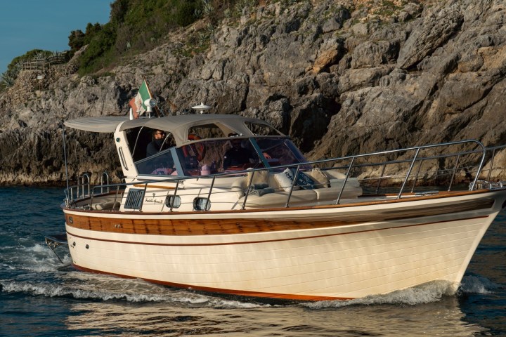 A boat with an Italian flag cruises near rocky cliffs on a sunny day.