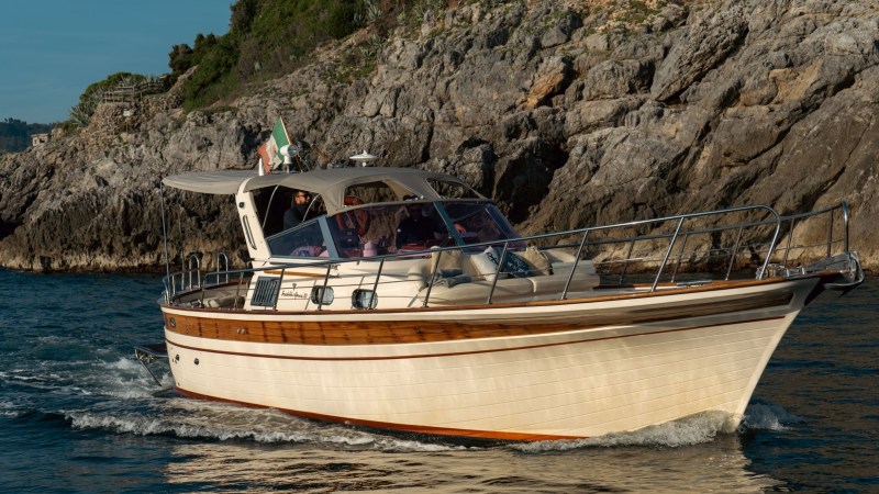 A boat with an Italian flag cruises near rocky cliffs on a sunny day.
