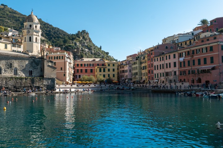 Colorful buildings by a harbor with a church tower and hills in the background under a clear sky.