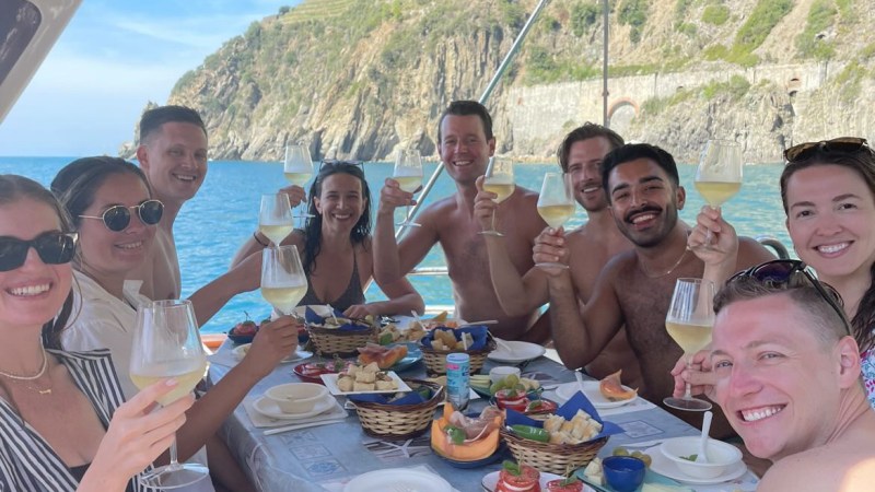 Group of people on a boat laughing and holding wine glasses, with food on the table and a coastal backdrop.