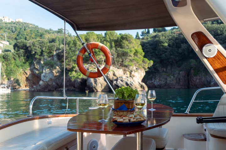 Boat deck with table, wine glasses, snacks, and view of rocky shoreline and trees.