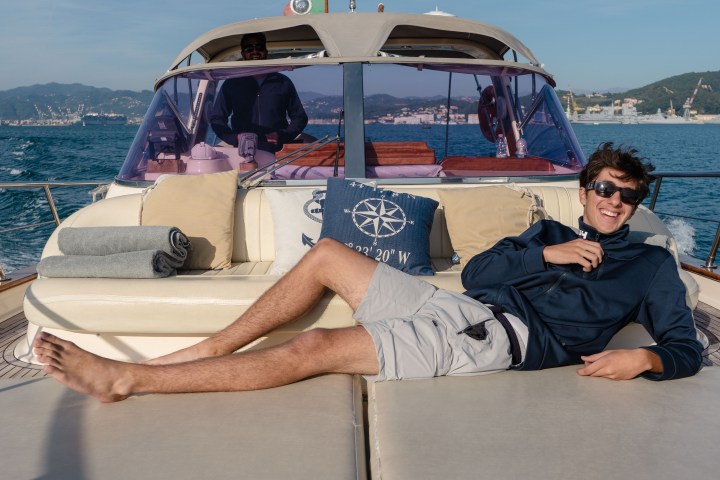 Person relaxing on a yacht's deck, wearing sunglasses, with scenic coastline in the background.