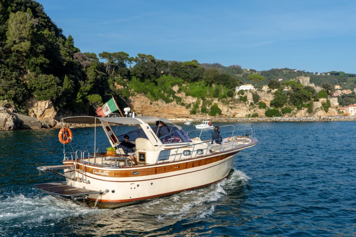 A boat sailing near a rocky coastline with trees and buildings in the background on a sunny day.