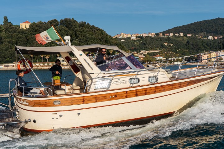 Motorboat cruising on water with three people and Italian flag, hillside town in background.