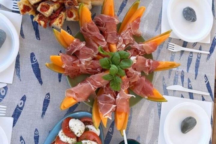 Italian antipasto with prosciutto, melon, caprese salad, cheese, and focaccia on a fish-patterned tablecloth.