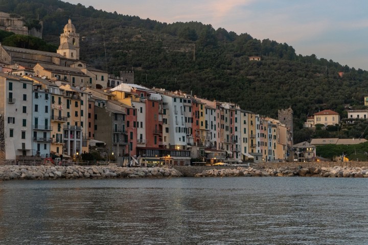 Colorful buildings along a waterfront with a hillside and church in the background at dusk.