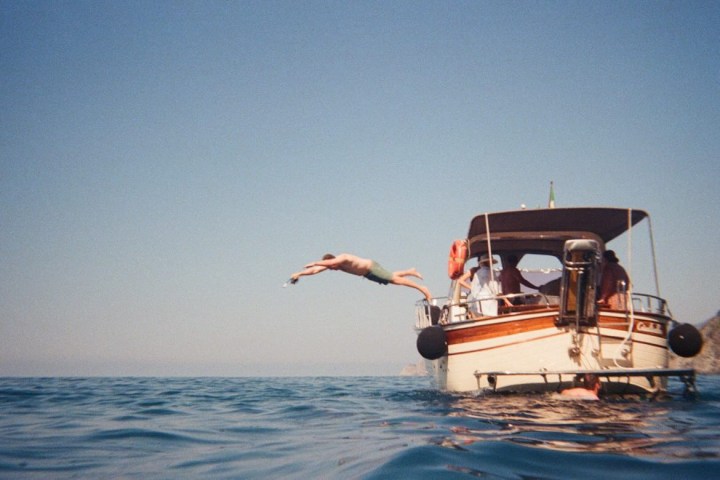 Person diving off a small boat into the ocean under a clear blue sky.