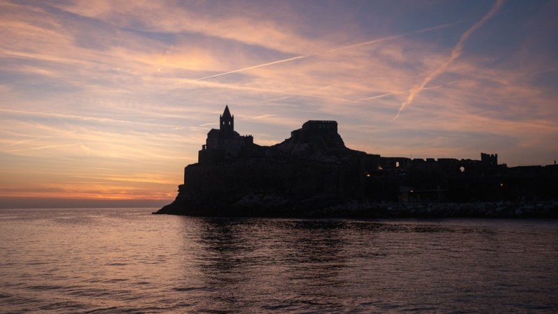 Silhouette of a fortress against a colorful sunset over a calm sea.