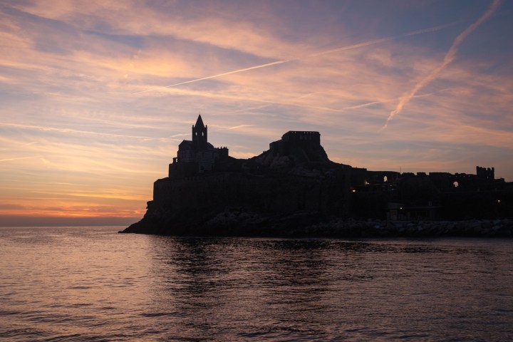 Silhouette of a coastal castle at sunset with colorful sky and calm sea.
