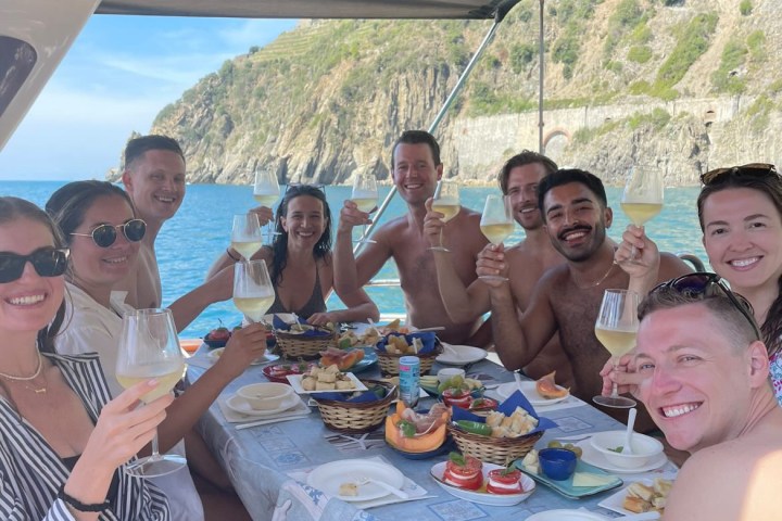 Group of people on a boat enjoying a meal and drinks, with a rocky coastline in the background.