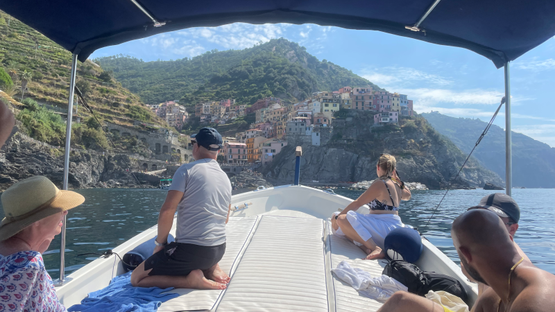 People on a boat approaching a coastal village with colorful houses on a hillside in the background.