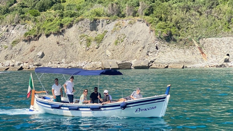 Group of people on a small boat with canopy near a rocky, green hillside.
