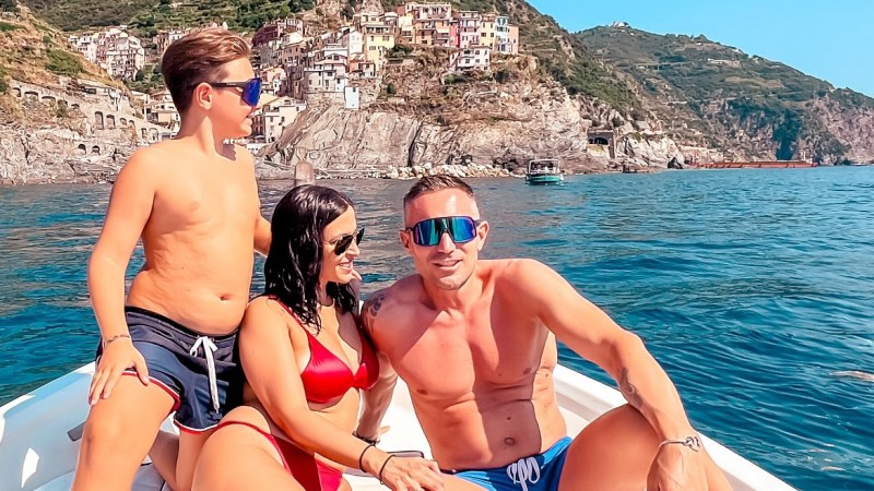 Three people on a boat with Manarola cliffs in the background under clear blue sky.