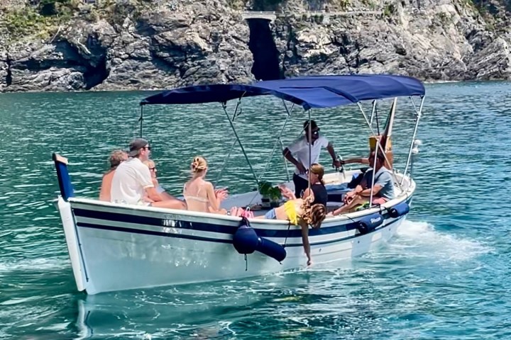 People relaxing on a boat with a blue canopy, near rocky cliffs and calm water.