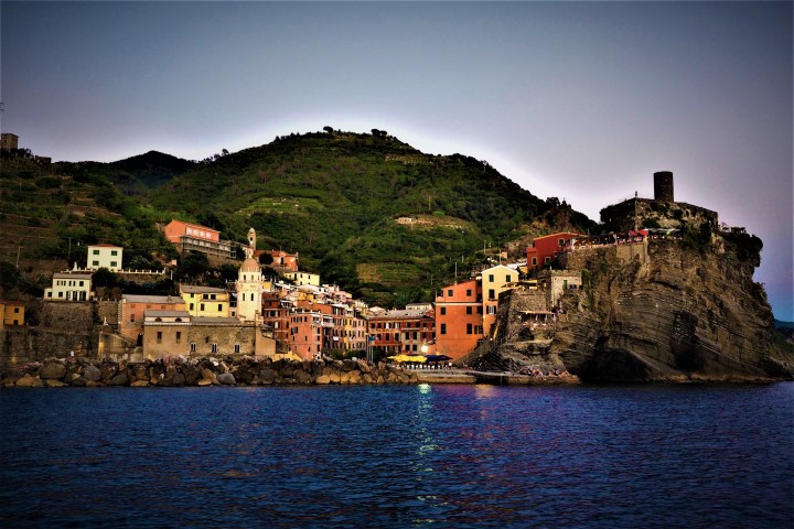 Scenic coastal village with colorful buildings, hillside, and tower by the sea at sunset.