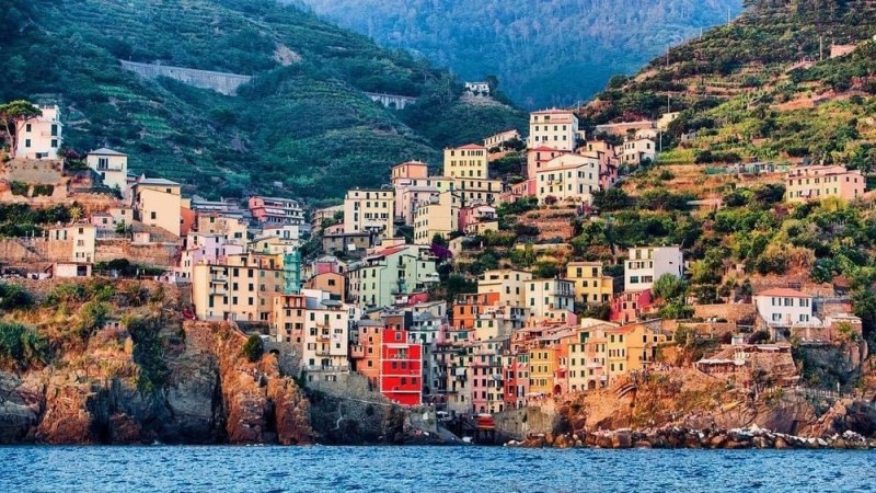 Colorful village on a hillside with sea in the foreground and mountains in the background.