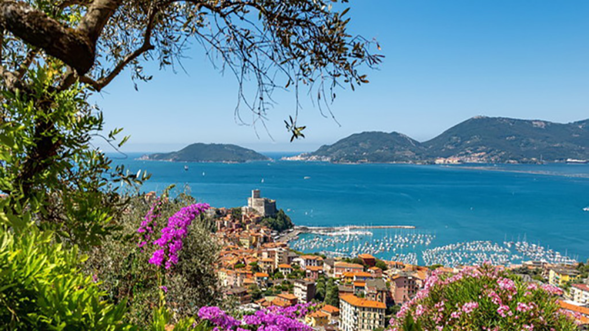 Coastal town with colorful flowers, sea, boats, and mountains under a clear blue sky.