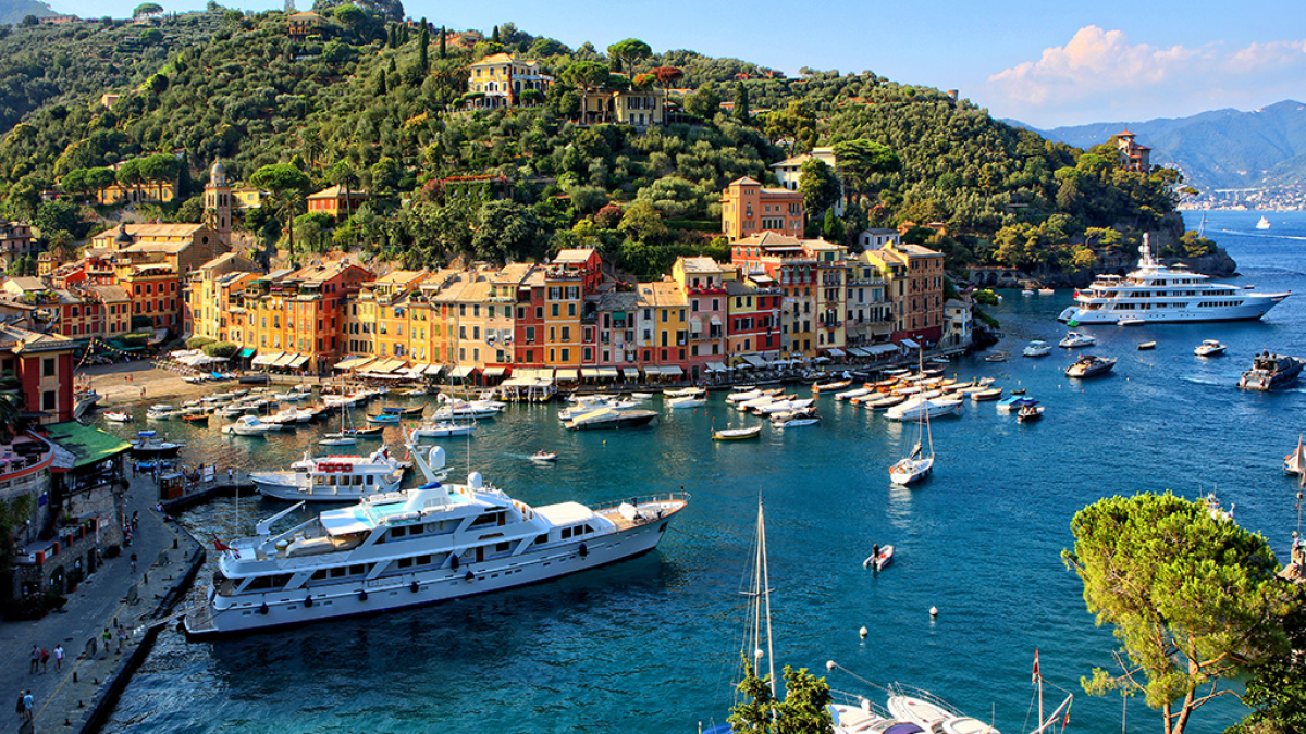 Colorful coastal town with yachts in a harbor and lush hillside backdrop.