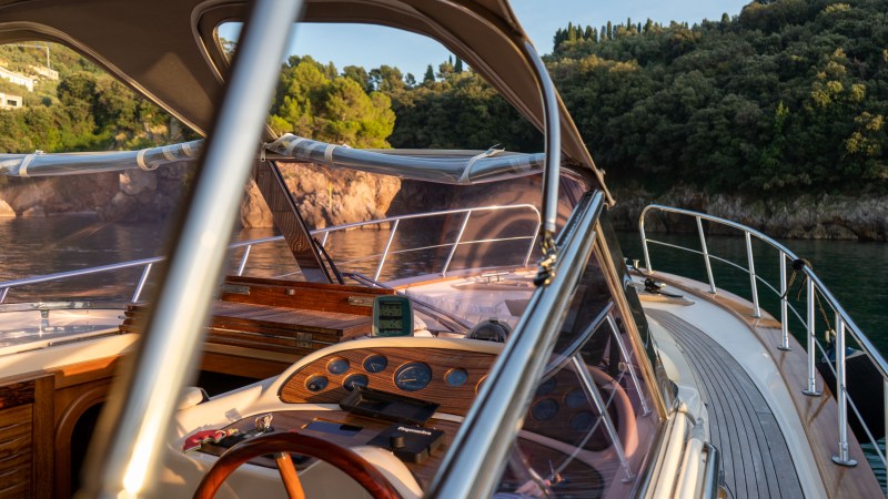 Boat cockpit with wooden dashboard, gauges, and view of trees and cliffs.