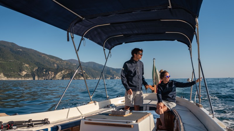 Two people in jackets on a boat with canopy, one pointing toward scenic coastline.