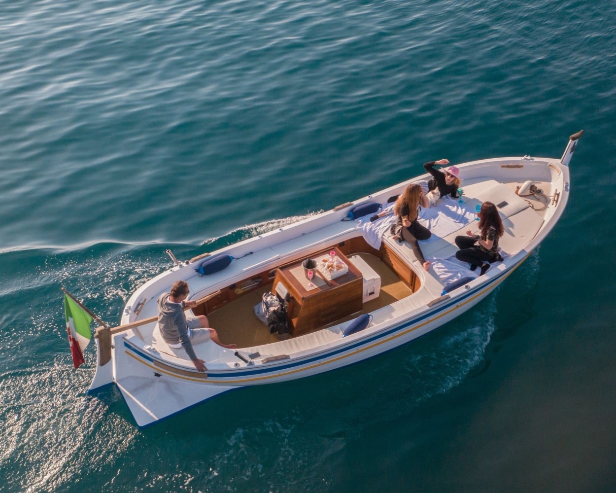 Four people relaxing on a small boat on calm water, with an Italian flag at the stern.